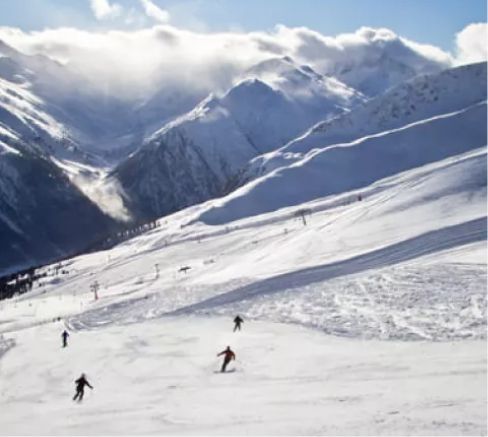 Mehrere Skifahrer fahren auf einem verschneiten Berg mit schneebedeckten Gipfeln im Hintergrund.